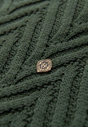 Strikket sweater i skovgrøn med et tekstureret mønster. Har et guldtonet badge med et indviklet design nær midten.