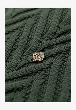 Strikket sweater i skovgrøn med et tekstureret mønster. Har et guldtonet badge med et indviklet design nær midten.