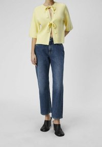 Cardigan giallo lavorato a maglia con scollo rotondo e fiocchi anteriori, abbinato a jeans in denim blu a vita alta e gamba dritta, insieme a mule nere con tacco.