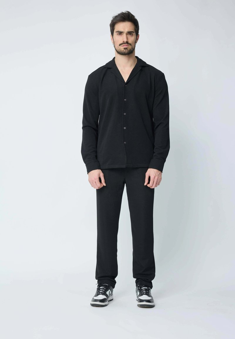 Chemise noire à manches longues avec un devant à boutons et un col, associée à un pantalon noir. Le modèle porte des baskets noires et argentées. Texture de tissu lisse.