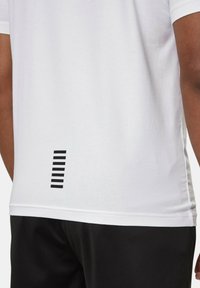 T-shirt bianco in cotone con maniche corte e un motivo a strisce orizzontali nere sul retro vicino all'orlo. Sono visibili anche pantaloni corti neri.