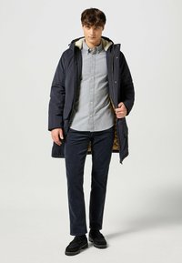 Abrigo largo navy con capucha forrada de pelo sintético, diseño acolchado y cierre de botón. Lleva puesto sobre una camisa gris de botones y pantalones oscuros. Zapatos negros.