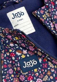 Marineblauwe bloemenjas met roze, gele en witte patronen, voorzien van een blauw etiket, ritsluiting en een fleece binnenvoering. Maattag zichtbaar.