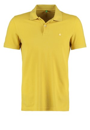 Mies keltainen poolopaita, mustat housut, valkoiset slip-on-kengät ja musta reppu selässään kävelee yksiväristä vaaleaa taustaa vasten.