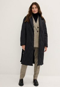 Cappotto lungo trapuntato nero con bottoni e superficie testurizzata, indossato sopra un completo doppiopetto a quadri e un dolcevita nero. Scarpe nere.