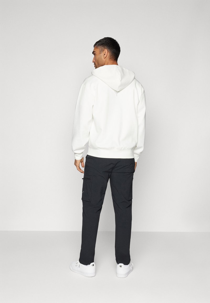 Sweatshirt à capuche blanche avec des poignets et un ourlet côtelés, associé à un pantalon cargo noir doté de plusieurs poches et des baskets blanches.
