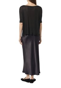 Czarna przezroczysta bluzka z krótkimi rękawami, nałożona na ciemną satynową maxi spódnicę. Czarne płaskie buty z ozdobnym zapięciem.