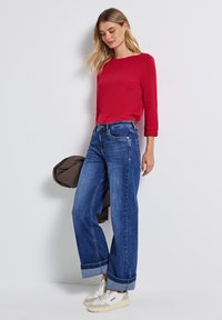 Červený dlouhý rukáv, široké modré džíny s ohrnutými lemy a bílé tenisky s béžovými detaily. Casual oblečení na modelce.