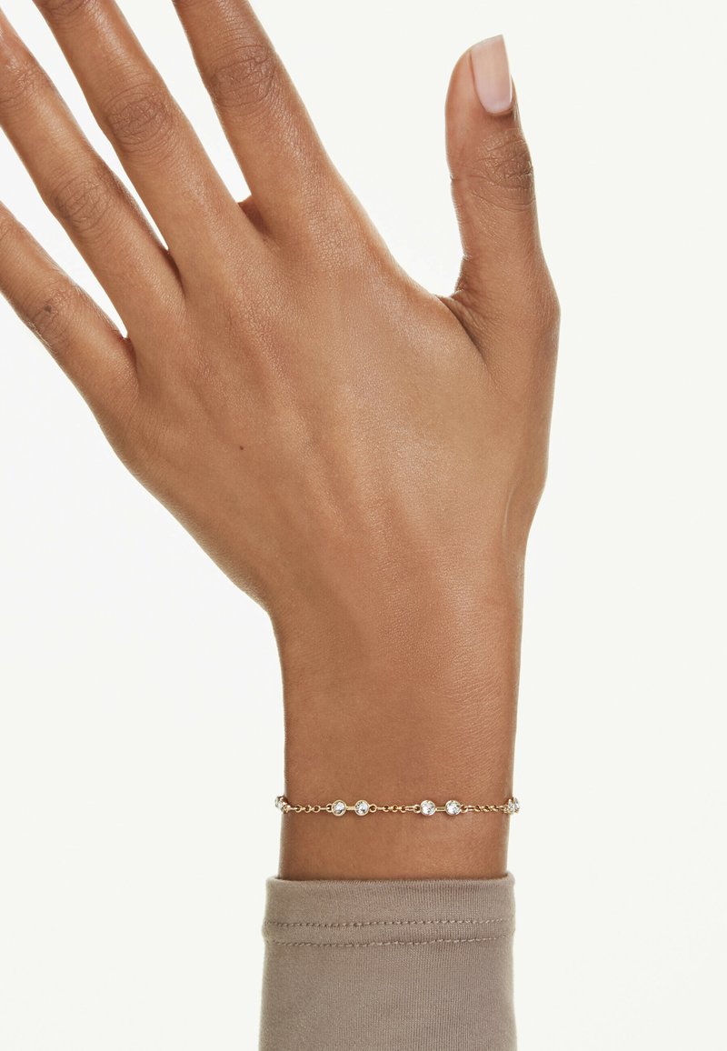 Guldtonad kedjearmband med flera klara, ovala stenar i en fin design. Bärs på en neutralt färgad ärm.