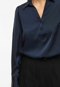 Tumši zila satīna blūze ar V veida izgriezumu, pogu lentu un garām piedurknēm. Kombinēta ar melnām biksēm, kurām ir piegriezums un jostas cilpas.