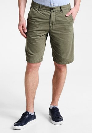 Homme portant un short olive jusqu'aux genoux, une chemise bleu clair rentrée, des chaussettes blanches à la cheville et des baskets basses noires.