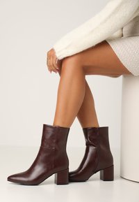 Stivaletti in pelle marrone scuro con finitura lucida, punta affusolata e tacco a blocco. Presentano una cerniera laterale e un branding minimale.