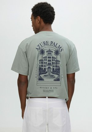 Hombre con camiseta verde claro con un gráfico detallado de un edificio de resort, palmeras y una fuente, combinado con pantalones blancos.