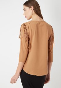 Blusa marrón con mangas tres cuartos, que presenta detalles de flecos a lo largo de los hombros. Tejido suave, dobladillo curvado y diseño sencillo.