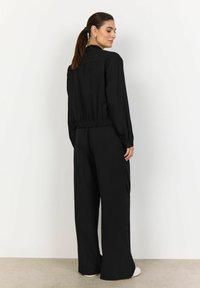 Jumpsuit nero con maniche lunghe, vita elastica e gambe larghe. Tessuto liscio con una sottile texture, progettato per una vestibilità rilassata.