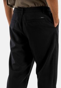 Vista posteriore di una persona che indossa pantaloni neri con tasche posteriori e mani nelle tasche laterali su sfondo semplice.