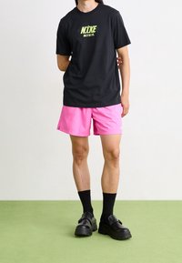 Musta T-paita vihreällä "NIKE"-logolla, yhdistettynä vaaleanpunaisiin shortseihin, mustiin sukkiin ja tukeviin mustiin kenkiin vihreällä lattiapinnalla. Yksinkertainen muotoilu, rento istuvuus.