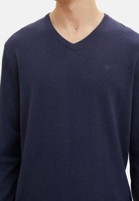 Marineblauer V-Ausschnitt-Pullover aus weichem Strickstoff. Verfügt über lange Ärmel und ein dezentes Logo auf der Brust, das eine glatte Textur bietet.