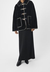 Zwarte bontjas met een grote kraag, voorzien van togglesluitingen, twee voorzakken en contrasterende stiksels. Gemaakt over een lange zwarte rok.