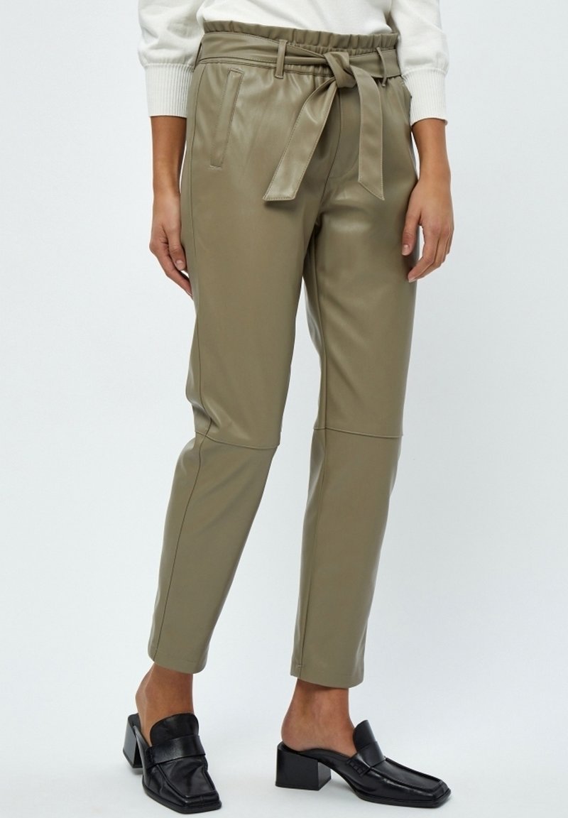 Femme portant un pantalon en cuir vert olive taille haute avec une ceinture nouée, associé à des mules à talons noires et un haut blanc à manches longues.