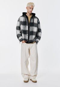 Karirana jakna u crno-bijeloj boji, nošena preko crne trenirke s kapuljačom, uparena s kremastim širokim hlačama i bež tenisicama, na jednobojnoj pozadini.