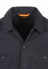 Donkerblauw button-up shirt met een puntige kraag, twee borstzakken en een oranje logolabel aan de binnenkant van de kraag. Vlotte textuur, klassiek ontwerp.