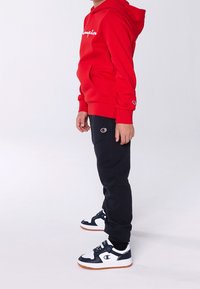 Sudadera roja con logo blanco, pantalones deportivos negros con logo y zapatillas en blanco y negro con suelas de goma. Atuendo atlético casual.