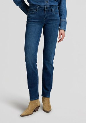 Person iført slanke mørkeblå jeans, en matchende denimskjorte og tanfarvede ruskindsstøvler, der står mod en ensartet baggrund.