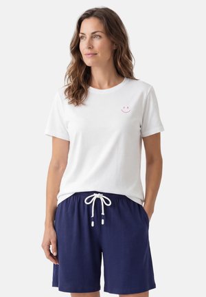 Kobieta o falowanych brązowych włosach, ubrana w białą koszulkę z małą różową buźką na piersi oraz granatowe szorty z białym sznurkiem.