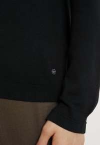 Zwarte gebreide trui met geribbelde manchetten, voorzien van een klein rond logo-label. De textuur is glad; het ontwerp is nauwsluitend.