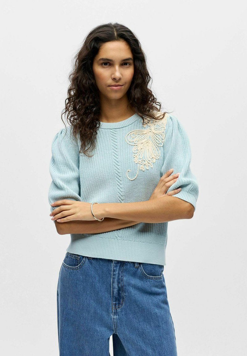 Pull en maille bleu clair avec manches courtes bouffantes, orné d'une broderie florale crème délicate sur l'épaule gauche. Associé à un jean en denim.