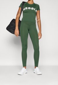 Zelený sportovní outfit se zkráceným rukávem s logem "NEBBIA", odpovídající legíny a bílé sportovní boty. Obsahuje černou sportovní tašku.