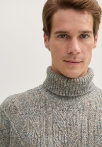 Hellegrauer Strickpullover mit hohem Kragen, der strukturierte Muster und dezente blaue Flecken im gesamten Stoff aufweist.