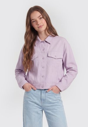 Giovane donna con capelli lunghi che indossa una giacca viola chiaro con bottoni e jeans a vita alta azzurri, mani nelle tasche, sfondo neutro.