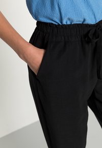 Czarne bawełniane spodnie z marszczonym paskiem i sznurkiem do wiązania. Po bokach na biodrach widoczne są kieszenie, a materiał wydaje się gładki.