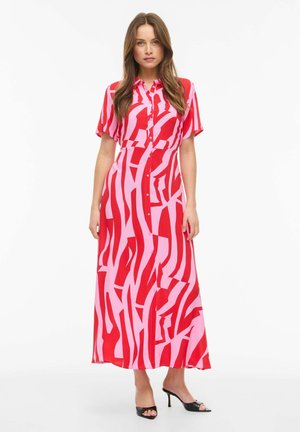 Kvinde iført en maxikjole med krave, korte ærmer og knapper foran med et abstrakt rødt og pink mønster, stående med sorte sandaler med hæl.