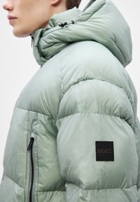 Ανοιχτό πράσινο μπουφάν με κουκούλα, με τσέπη με φερμουάρ και μαύρη τετράγωνη ετικέτα στο μανίκι με τη λέξη "BOSS" κεντημ�ένη σε χάλκινο χρώμα.