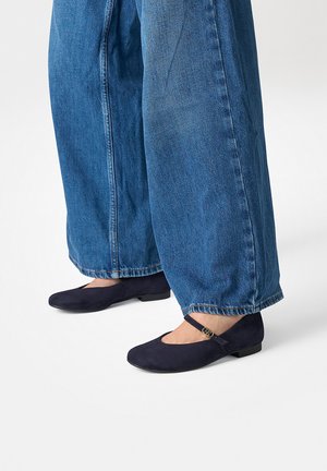 Person trägt weite blaue Jeans und dunkle navyfarbene Mary Jane Ballerinas mit einer goldenen Schnalle und steht auf einer weißen Oberfläche.