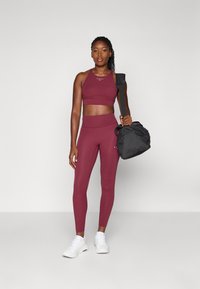 Burgunderfarbener Sport-Crop-Top und Leggings aus dehnbarem Material, kombiniert mit einer schwarzen Sporttasche. Weiße Sneakers runden das Outfit ab.