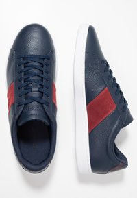 Marinblå lädersneakers med en röd mockadetalj, vit gummisula och texturerad ovandel med kontrasterande snörning.