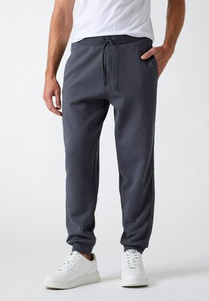 Uomo che indossa pantaloni jogger grigio scuro con vita con coulisse e sneakers bianche, in piedi con una mano in tasca su sfondo bianco.