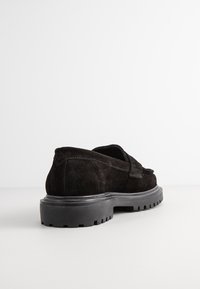 Mocassin en daim noir avec semelle crantée épaisse, détails cousus, et un petit talon, vu de l'arrière sur un fond blanc.