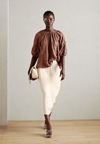 Braune, strukturierte Bluse mit Puffärmeln; weiße, plissierte Midirock; klare Brille; hält eine beige Handtasche; steht auf einem Holzboden.