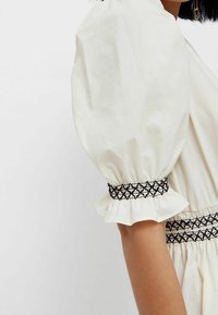 Hvid kjole med pufærmer, volangkant og sorte smock-detaljer ved manchetterne og i taljen. Glat stof, afslappet pasform.