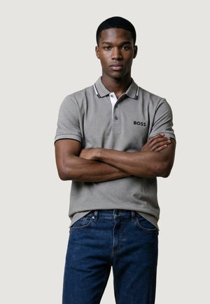 Muž se zkříženýma rukama, oblečený v šedém poloshirtu BOSS a modrých džínách, stojící před jednoduchým světle šedým pozadím.