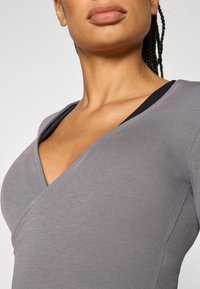 Γκρι ριπ τοπ με βαθύ V-λαιμόκοψη, με μαύρες λεπτομέρειες κατά μήκος του λαιμού. Το ύφασμα φαίνεται λείο και εφαρμοστό.