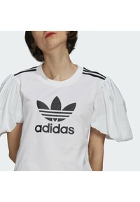Camisa blanca de manga corta con mangas abullonadas y logo negro de Adidas. Presenta tres rayas negras en los hombros. Material de algodón con textura suave.
