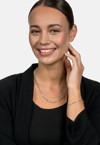 Silver jewelry consisting of layered necklaces with delicate links and small charms, paired with a matching bracelet, worn against a black top.
