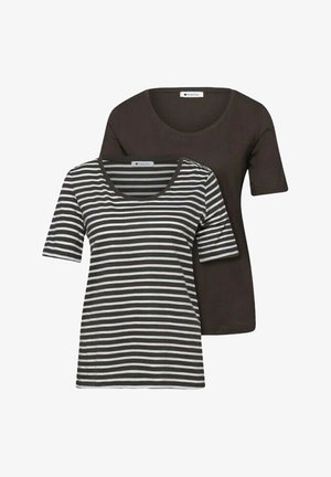 Due donne con jeans blu casual e t-shirt stanno una a fianco all'altra contro un muro bianco; una tiene una giacca blu.