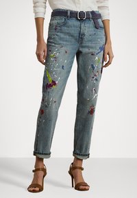 Gaiši zili džinsi ar brīvu piegriezumu, kuriem ir krāsaini krāsas traipi. Cieši sasieti piedurknes un pītā josta izceļ dizainu.